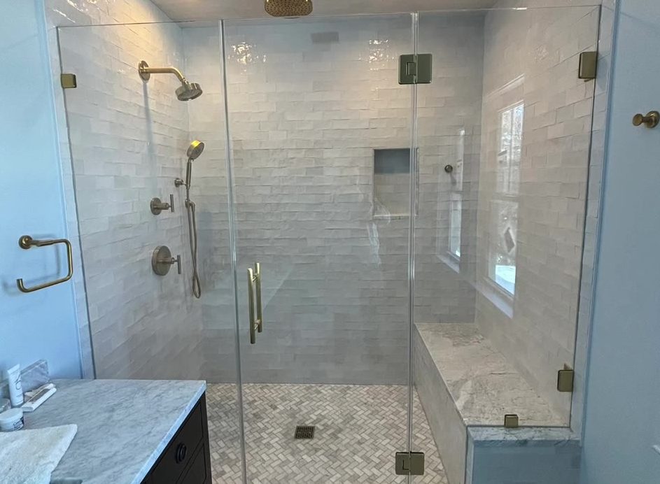 The same shower after: a frameless glass door and panel with brass-finish hinges and pull, bench and window inside the enclosure