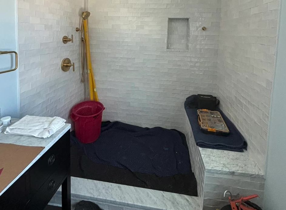 Shower alcove mid-construction before glass: white tile, a covered bench, tools and a bucket on the floor, brass fixtures