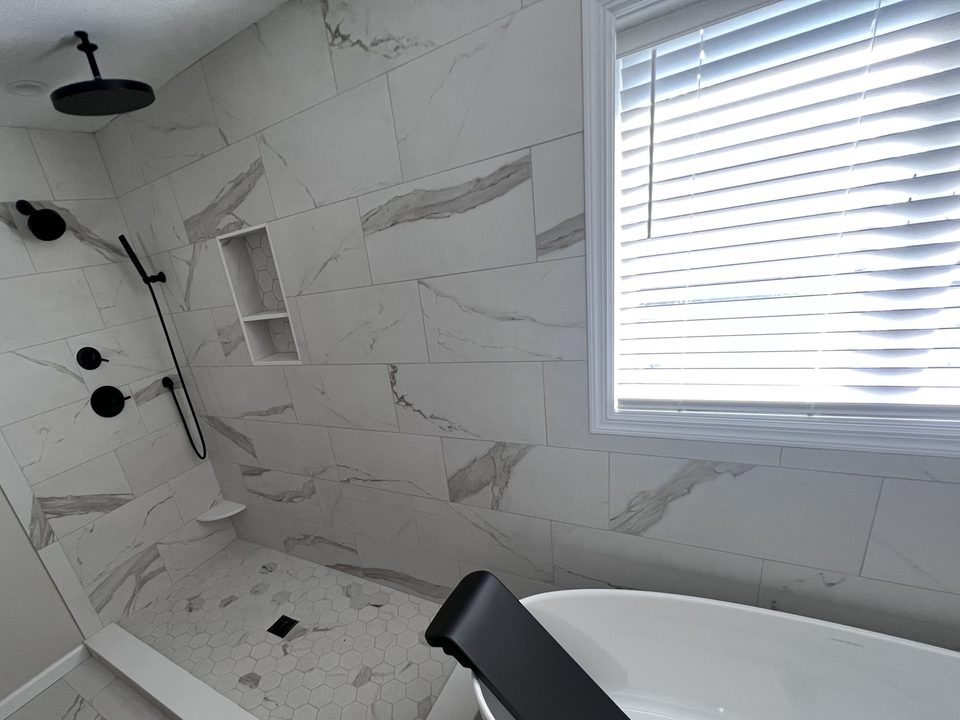 Corner shower before glass: marble-look tile, black rain head and fixtures, a hex-tile floor and a freestanding tub alongside