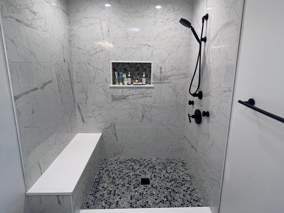 Tiled walk-in shower alcove before glass: marble-look walls, a white bench, black rain head and handheld, penny mosaic floor