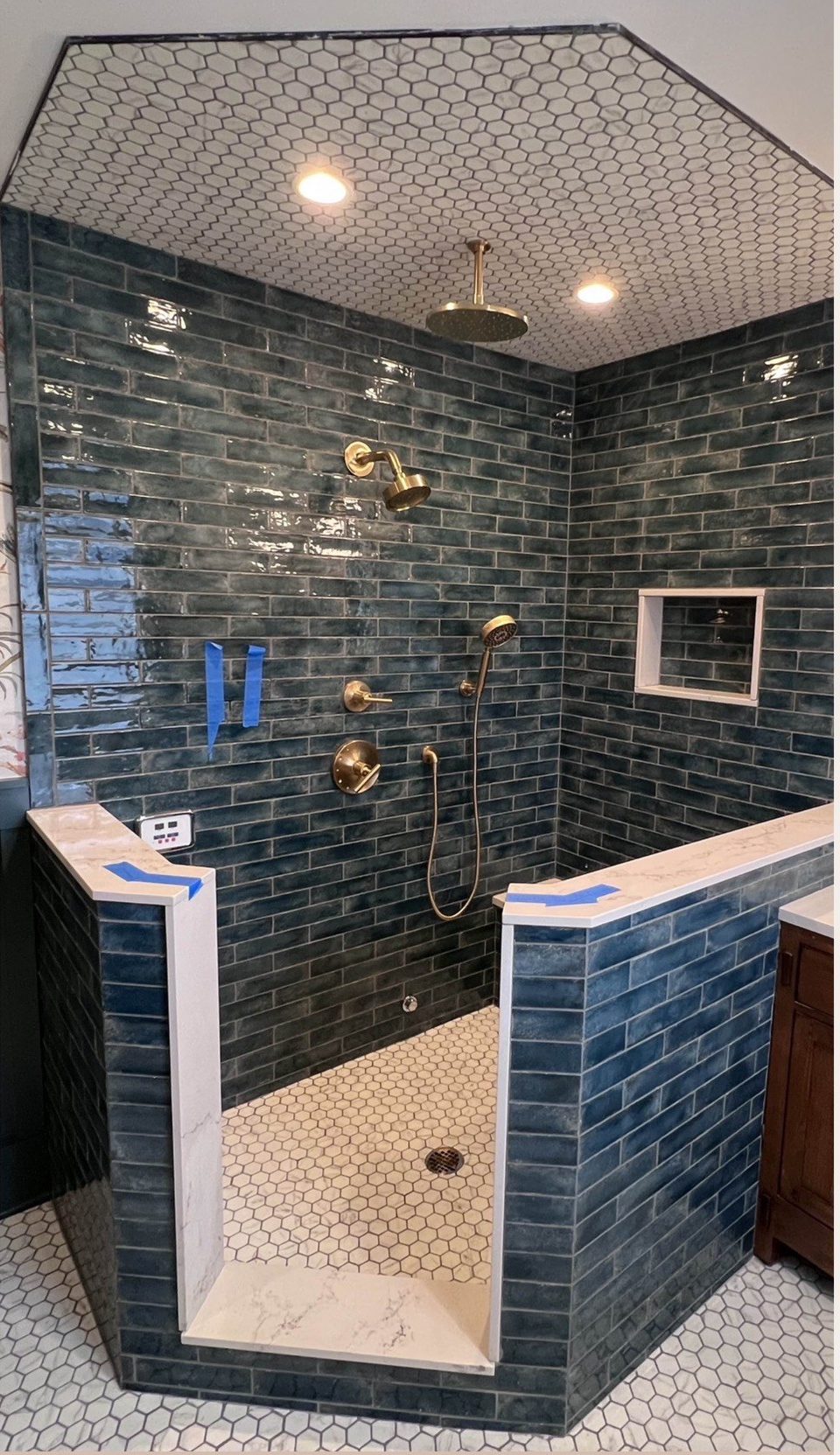 Corner shower before glass: blue-green subway tile, brass rain head and fixtures, low tiled knee walls, hex-tile ceiling