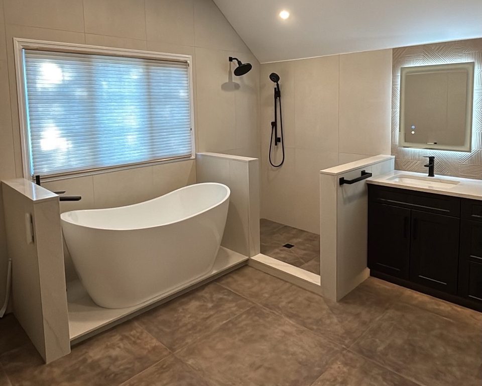 Bathroom before glass: a freestanding tub and a bare tiled walk-in shower between two low knee walls, black shower fixtures, dark vanity at right