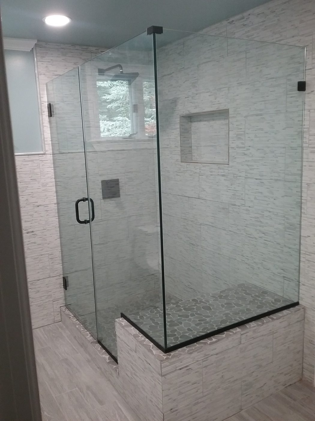 Corner frameless enclosure with black clips, hinged off the wall, on a pebble floor with a tiled half wall