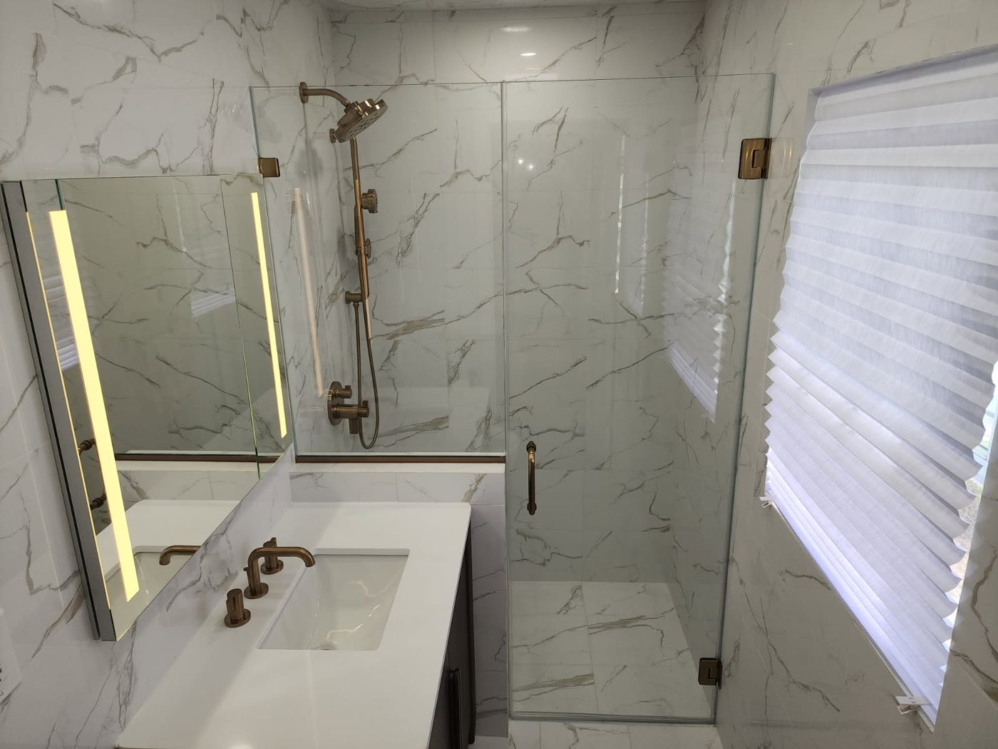 Frameless shower door in marble with brass fixtures beside a lighted vanity mirror